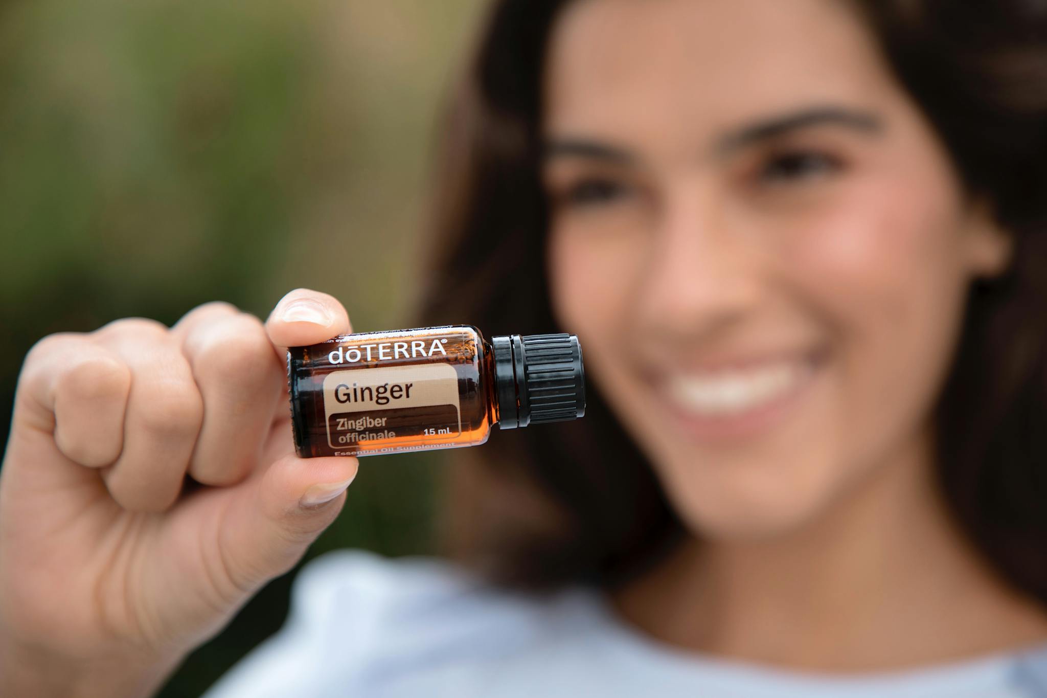 A smiling woman holds a doterra ginger essential oil bottle outdoors, showcasing natural wellness products.